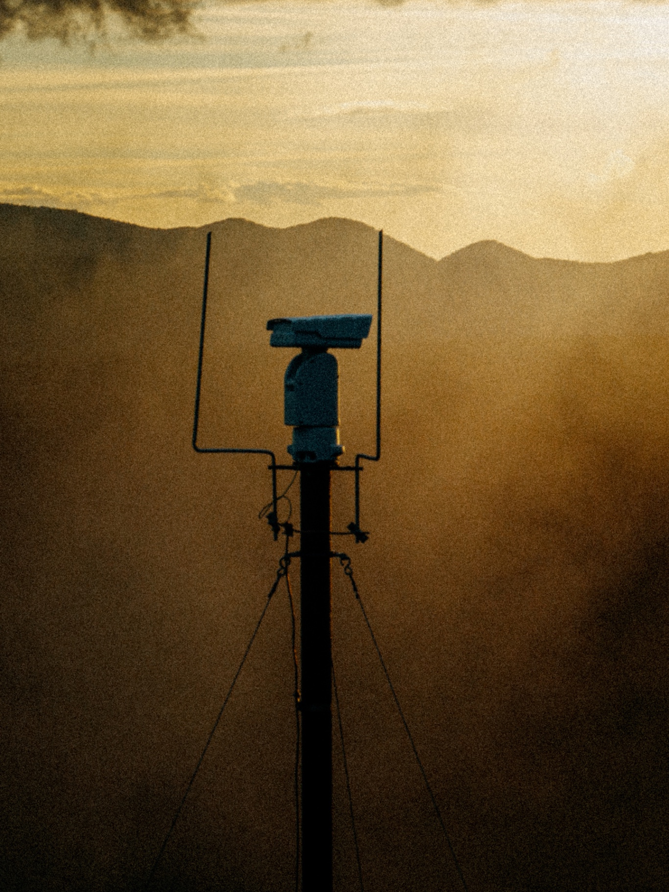 Close-up of a LAST-Fire pan-tilt camera head on a mast against a hazy golden sunset and mountains